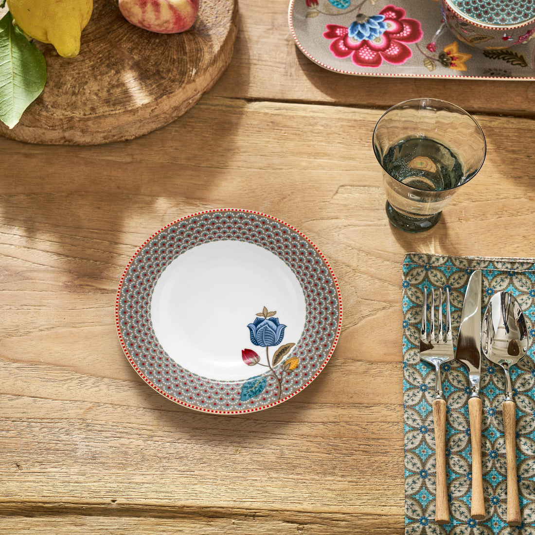 Dining table setting with floral plates, cutlery, and decorative items on a wooden surface.