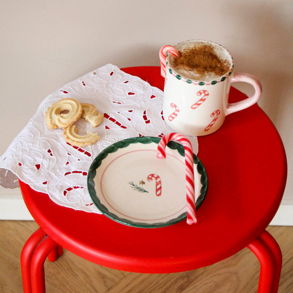 Rice kerst gebaksbordje met candy cane motief en een zuurstok naast een bijpassende mok op een rode kruk.